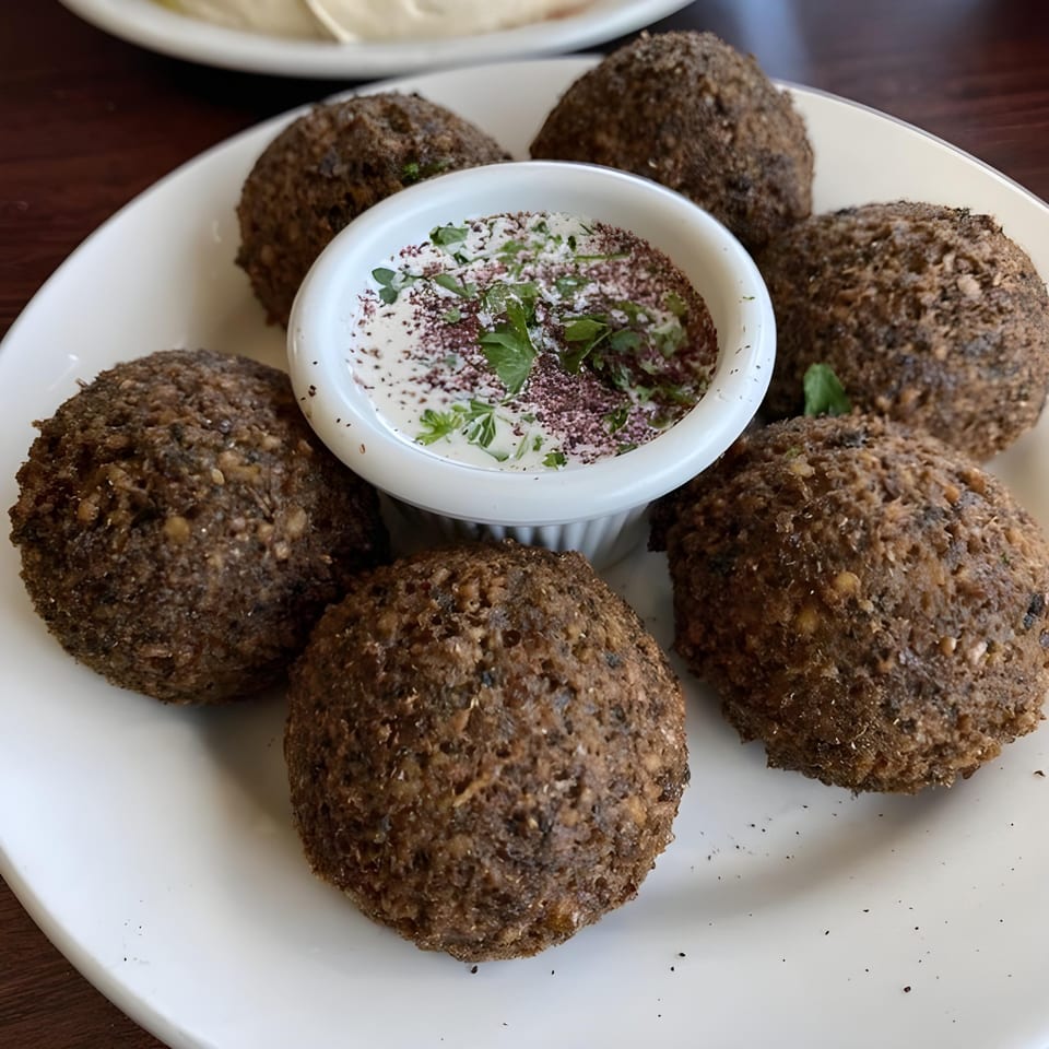 Six crispy falafel balls surround a white bowl of creamy sauce, garnished with sumac and parsley.