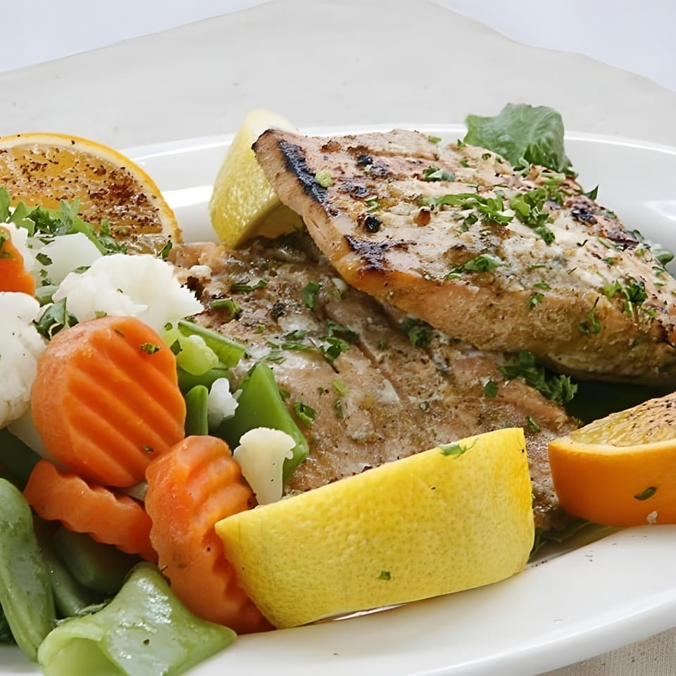 Grilled fish fillets with steamed carrots, green beans, cauliflower, and lemon wedges on a white plate.